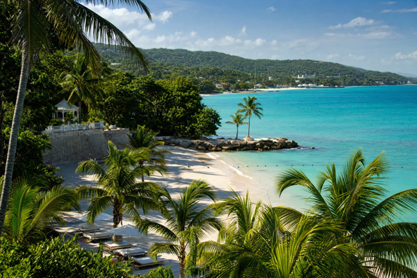 A panoramic view of a Jamaican beach with turquoise water, palm trees, and lush mountains in the background.